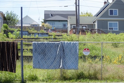 A residential area with two houses in the background. In the foreground, there are items hanging on a chain-link fence, including a patterned blanket and a dark-colored fabric. A 'No Trespassing' sign is attached to the fence. The scene includes a grassy yard with some overgrown plants.