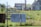 A residential area with two houses in the background. In the foreground, there are items hanging on a chain-link fence, including a patterned blanket and a dark-colored fabric. A 'No Trespassing' sign is attached to the fence. The scene includes a grassy yard with some overgrown plants.
