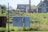 A residential area with two houses in the background. In the foreground, there are items hanging on a chain-link fence, including a patterned blanket and a dark-colored fabric. A 'No Trespassing' sign is attached to the fence. The scene includes a grassy yard with some overgrown plants.