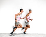 Two men are running alongside each other against a plain white background. They are wearing matching athletic outfits, consisting of white sleeveless shirts with pink and grey stripes, grey shorts, and black running shoes with black socks.