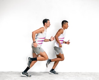 Two men are running alongside each other against a plain white background. They are wearing matching athletic outfits, consisting of white sleeveless shirts with pink and grey stripes, grey shorts, and black running shoes with black socks.