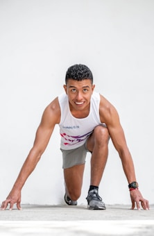 A young man is poised in a starting position for a race. He is wearing a white tank top with the word 'RUNNER' and gray shorts. His face shows determination and focus, and he wears a watch on his wrist. The background is plain and white, emphasizing the subject.