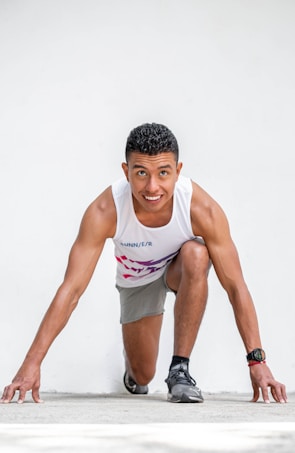A young man is poised in a starting position for a race. He is wearing a white tank top with the word 'RUNNER' and gray shorts. His face shows determination and focus, and he wears a watch on his wrist. The background is plain and white, emphasizing the subject.