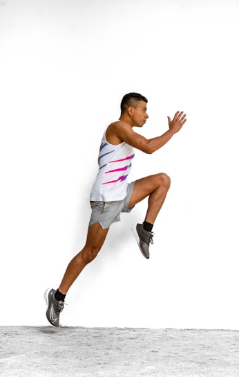 A man is portrayed in mid-air, performing a dynamic running pose against a plain white background. He wears a sleeveless sports top with blue and pink accents, along with gray shorts and black athletic shoes.
