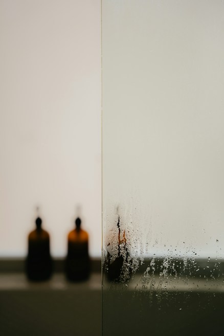 A minimalistic and serene composition featuring a frosted glass panel, with water droplets and condensation on the surface. In the background, two blurred brown glass bottles with pump tops are visible, adding a touch of elegance and simplicity.