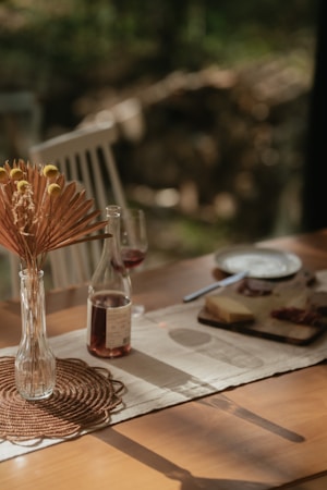A wooden table is set with a glass vase containing dried flowers, a bottle of wine, and two wine glasses. A wooden cheese board with cheese and charcuterie is placed next to a knife. Soft lighting casts gentle shadows across a woven mat and a beige table runner, with a blurred background of greenery.