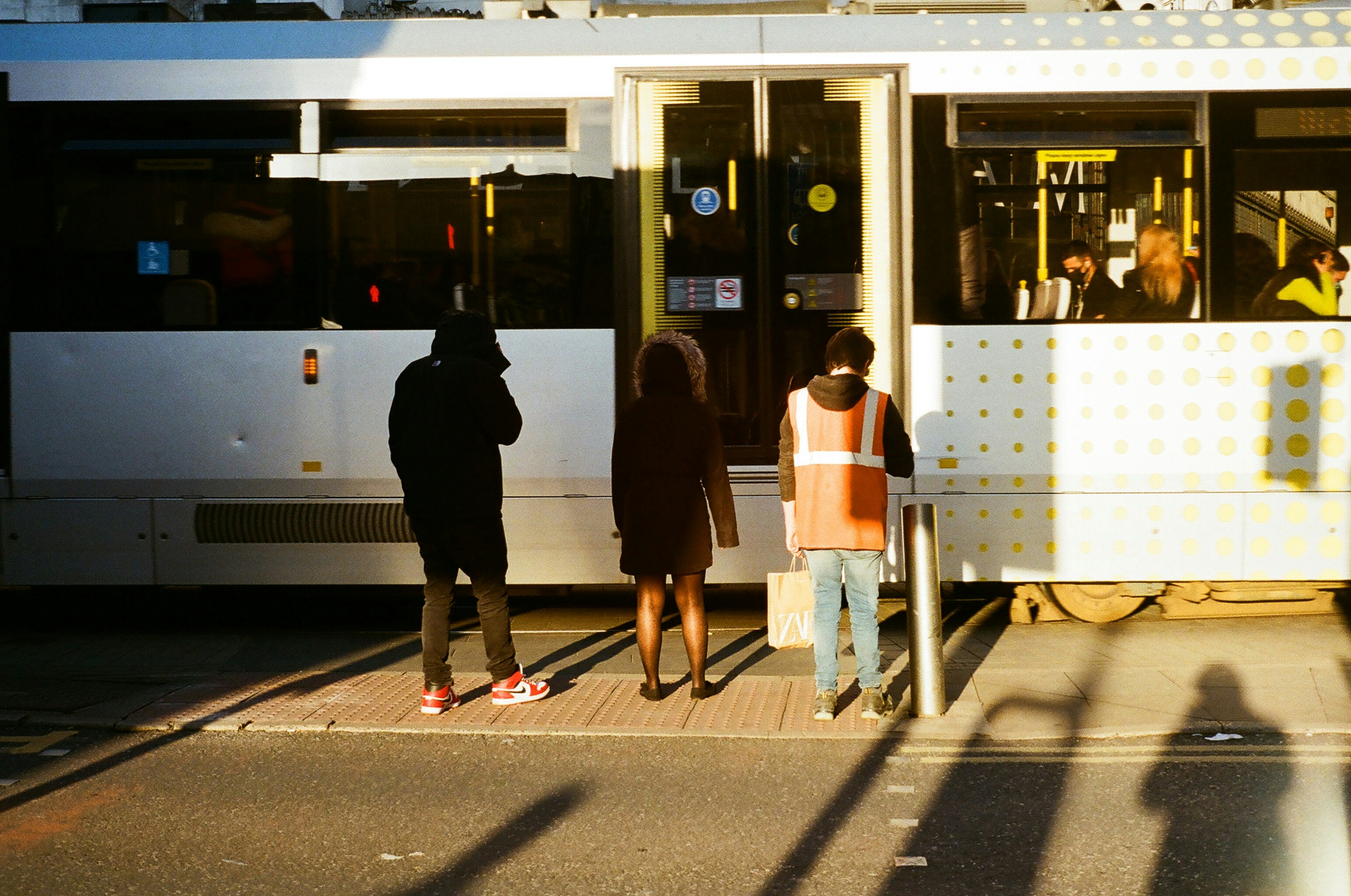 Landscape street photograph showing three individuals waiting for a Manchester Metrolink tram to drive past them.