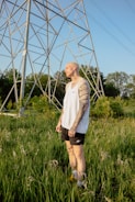 A tattooed person is standing in a field of tall green grass under a large metal structure resembling a power line tower. The individual is wearing a white sleeveless shirt and black shorts, with a contemplative expression on their face. The background features lush trees and a clear blue sky, creating a serene outdoor setting.