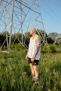 A tattooed person is standing in a field of tall green grass under a large metal structure resembling a power line tower. The individual is wearing a white sleeveless shirt and black shorts, with a contemplative expression on their face. The background features lush trees and a clear blue sky, creating a serene outdoor setting.