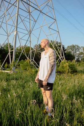 A tattooed person is standing in a field of tall green grass under a large metal structure resembling a power line tower. The individual is wearing a white sleeveless shirt and black shorts, with a contemplative expression on their face. The background features lush trees and a clear blue sky, creating a serene outdoor setting.