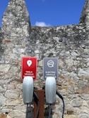Two electric vehicle charging stations with signs attached to a stone wall. One sign reads 'Tesla Vehicle Charging' and the other reads 'Electric Vehicle Charging.' The background features a rustic, weathered stone wall beneath a clear blue sky.