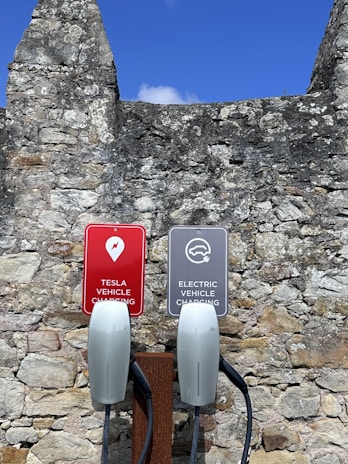 Two electric vehicle charging stations with signs attached to a stone wall. One sign reads 'Tesla Vehicle Charging' and the other reads 'Electric Vehicle Charging.' The background features a rustic, weathered stone wall beneath a clear blue sky.