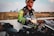 A person stands next to a motorcycle in an outdoor setting. The individual is wearing a leather jacket with green accents and a cap. A helmet with several logos, including one for Monster Energy, is placed on the motorcycle. In the background, a city skyline and an open field are visible.