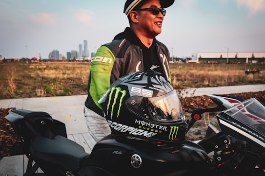 A person stands next to a motorcycle in an outdoor setting. The individual is wearing a leather jacket with green accents and a cap. A helmet with several logos, including one for Monster Energy, is placed on the motorcycle. In the background, a city skyline and an open field are visible.