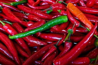 An assortment of vibrant peppers in varying shades, predominantly red with a few green and orange ones mixed in. The peppers are shiny and have a fresh appearance, piled closely together.