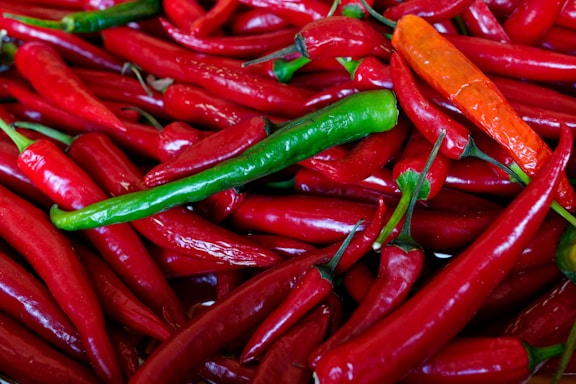 An assortment of vibrant peppers in varying shades, predominantly red with a few green and orange ones mixed in. The peppers are shiny and have a fresh appearance, piled closely together.