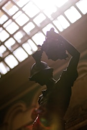 A silhouetted sculpture of a warrior holding a severed head, dramatically lit from above by sunlight streaming through a patterned glass ceiling. The details of the warrior's helmet and muscular form are highlighted against the bright backlight.