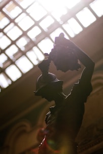 A silhouetted sculpture of a warrior holding a severed head, dramatically lit from above by sunlight streaming through a patterned glass ceiling. The details of the warrior's helmet and muscular form are highlighted against the bright backlight.