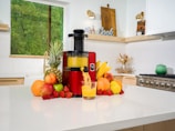 A modern kitchen with a white countertop has a red juicer machine in the center, surrounded by a variety of fruits including pineapples, bananas, apples, strawberries, and oranges. Fresh orange juice is being poured into a glass from the juicer. The background shows a wooden cutting board with the word 'Cook' on it, a window with a green view, and a green pot on a stove.