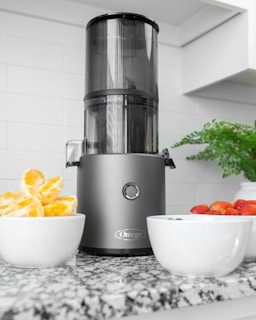 A modern kitchen setting features a sleek black and metallic juicer placed on a speckled granite countertop. To the left, a white bowl filled with segmented oranges adds a bright pop of color. On the right, another white bowl contains fresh strawberries and a few blueberries. In the background, white tiled walls and a potted green plant add to the clean, fresh look of the space.