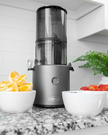 A modern kitchen setting features a sleek black and metallic juicer placed on a speckled granite countertop. To the left, a white bowl filled with segmented oranges adds a bright pop of color. On the right, another white bowl contains fresh strawberries and a few blueberries. In the background, white tiled walls and a potted green plant add to the clean, fresh look of the space.