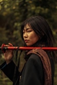 A person with long dark hair holds a red staff over their shoulder while standing in a natural, leafy setting. They wear a dark coat and an intricately patterned scarf, with a serious and contemplative expression.
