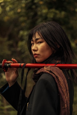 A person with long dark hair holds a red staff over their shoulder while standing in a natural, leafy setting. They wear a dark coat and an intricately patterned scarf, with a serious and contemplative expression.