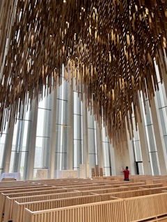 An interior view of a modern chapel with tall vertical windows and wooden pews. The ceiling features an intricate installation of hanging wooden rods creating an artistic pattern. A person in a red shirt is standing among the pews, taking a photo or video.