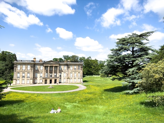 A large, stately mansion with classical architecture is situated in the middle of a well-manicured green lawn with yellow flowers. The sky is bright blue with scattered white clouds. In the foreground, two people are sitting on the grass, enjoying the serene landscape. A massive tree with lush foliage is positioned to the right, adding to the idyllic setting. Surrounding the estate, there are more trees, indicating a lush, expansive garden or park.