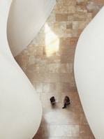 Aerial view of a tiled floor with two people walking across it, surrounded by large, smooth, white architectural curves. The light creates a bright reflection on the floor, adding a sense of warmth.