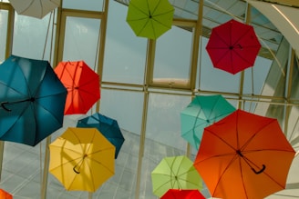 A collection of colorful umbrellas is suspended from the ceiling inside a building, with a glass roof allowing natural light to filter through. The umbrellas vary in colors including red, blue, green, yellow, and orange.