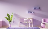 The image depicts a minimalist, modern interior with a focus on a soft pastel color scheme. A potted plant with large green leaves stands on the floor next to a light-colored desk and chair. Above the desk, two floating shelves hold small books and decorative items. A cozy pink armchair sits to the right, adjacent to the purple-toned wall. The entire setting is grounded by a purple rug that matches the pastel theme.