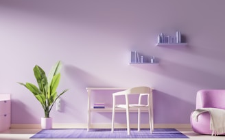 The image depicts a minimalist, modern interior with a focus on a soft pastel color scheme. A potted plant with large green leaves stands on the floor next to a light-colored desk and chair. Above the desk, two floating shelves hold small books and decorative items. A cozy pink armchair sits to the right, adjacent to the purple-toned wall. The entire setting is grounded by a purple rug that matches the pastel theme.