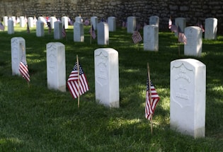 Several white gravestones are lined up in rows, each marked with a small American flag on well-maintained green grass. The background features a stone wall. Some of the gravestones are inscribed with the words 'Unknown U.S. Soldier.'