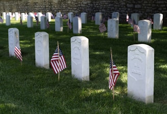 Several white gravestones are lined up in rows, each marked with a small American flag on well-maintained green grass. The background features a stone wall. Some of the gravestones are inscribed with the words 'Unknown U.S. Soldier.'