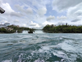 A vast and powerful waterfall cascades down rocky cliffs, surrounded by lush green trees and vegetation. A historical-looking building and other structures are perched on the cliffs, overlooking the turbulent waters. The sky above is filled with scattered clouds, creating a dramatic and picturesque landscape.