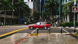 A red car is driving through an urban intersection surrounded by tall buildings and palm trees. The streets are lined with clear road markings, and a few pedestrians are visible on the sidewalk. Street signs and traffic lights are present, with one showing a red signal.