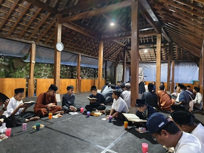 A group of people, mostly young men, are sitting cross-legged on the floor of a wooden pavilion. They are holding and reading books, likely engaged in a collective reading or study session. The pavilion has a high, exposed wooden roof and is illuminated by overhead lighting. There are beverages and snacks placed in front of some individuals, suggesting an informal or social gathering.