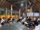 A group of people, mostly young men, are sitting cross-legged on the floor of a wooden pavilion. They are holding and reading books, likely engaged in a collective reading or study session. The pavilion has a high, exposed wooden roof and is illuminated by overhead lighting. There are beverages and snacks placed in front of some individuals, suggesting an informal or social gathering.