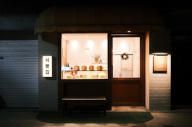 A small bakery is warmly lit from the inside, showcasing various loaves of bread in the window display. The exterior features an awning above the display window and door. A bench is placed in front of the display window. There are signs on both sides of the bakery door, one with lighting. Christmas-themed decor, such as a wreath, can also be seen on the door inside.