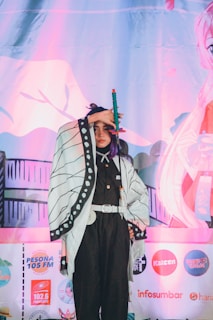 A person in a detailed costume stands in front of a colorful backdrop featuring various logos and anime-style artwork. They are holding a sword with a green handle and an orange tip, wearing a black outfit with a patterned white cloak.
