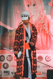 A person dressed in a colorful, traditional Japanese-inspired costume that includes a patterned robe and a sword. The individual is wearing a hat with a distinct black and white design. In the background, there is large artwork depicting an anime character with pink hues and cherry blossom motifs. Various logos and symbols are also visible in the lower portion of the image.