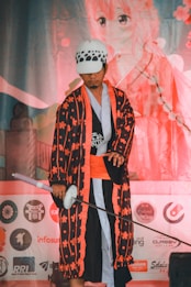 A person dressed in a colorful, traditional Japanese-inspired costume that includes a patterned robe and a sword. The individual is wearing a hat with a distinct black and white design. In the background, there is large artwork depicting an anime character with pink hues and cherry blossom motifs. Various logos and symbols are also visible in the lower portion of the image.