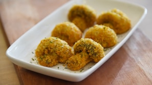 A white rectangular plate holds several golden-brown, breaded mushroom caps garnished with herbs, displayed on a wooden surface.