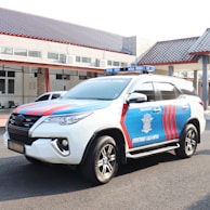 A marked police SUV with red and blue stripes is parked on a paved surface. The vehicle has a roof-mounted light bar and the words 'Direktorat Lalu Lintas' visible on the side. It is parked in front of a building with a tiled roof and several windows.
