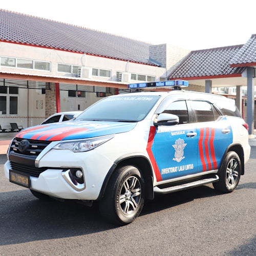 A marked police SUV with red and blue stripes is parked on a paved surface. The vehicle has a roof-mounted light bar and the words 'Direktorat Lalu Lintas' visible on the side. It is parked in front of a building with a tiled roof and several windows.