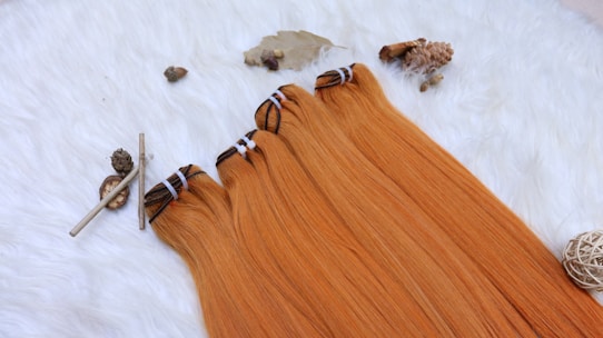 Four bundles of straight, orange hair extensions lying on a white, fluffy surface. Surrounding the hair are natural elements including pine cones, twigs, and a woven decorative ball, creating an organic, rustic atmosphere.