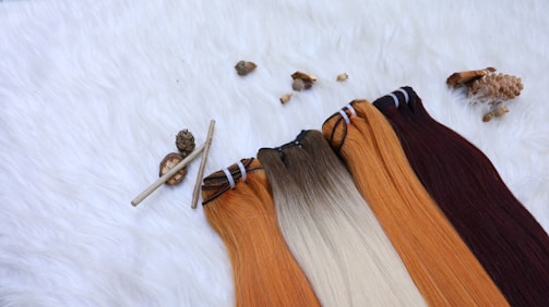 Several hair extensions in varying shades from orange to brown are laid out on a furry white surface. Natural elements like small pine cones and sticks add a rustic touch to the arrangement.