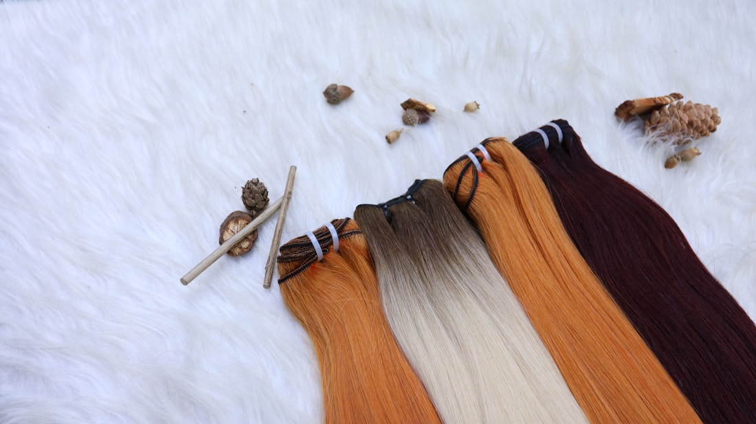 Several hair extensions in varying shades from orange to brown are laid out on a furry white surface. Natural elements like small pine cones and sticks add a rustic touch to the arrangement.