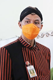 A person wearing a traditional black and red striped outfit and an orange face mask stands facing forward. The outfit includes a headpiece, and the person is wearing an ID badge and a button on their chest.